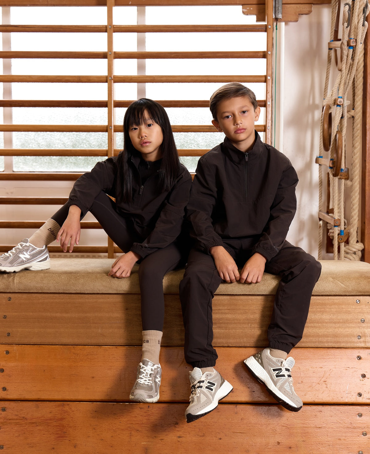 Two children in dark matching tracksuits sitting on a gym bench